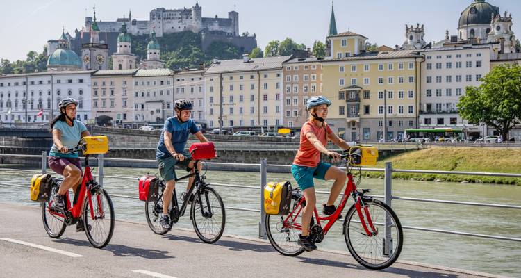 Trois cyclistes roulent le long de la rivière Salzach avec l'horizon baroque de Salzbourg et sa forteresse bien en vue.
