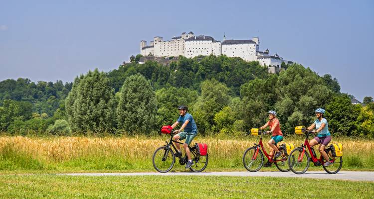 Des cyclistes parcourent un sentier de campagne avec des champs de blé devant l'imposante forteresse perchée de Salzbourg.
