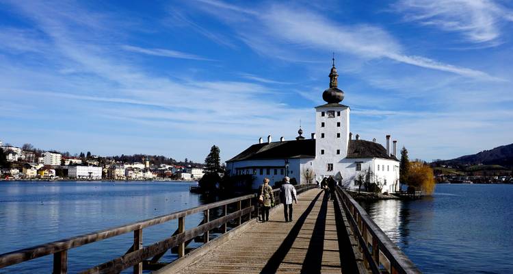 A lakeside castle with a wooden walkway leading to it, with people walking along the bridge.