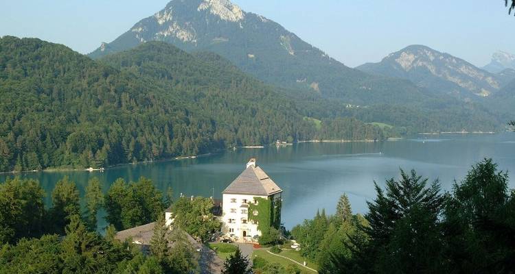 Un château au bord d'un lac avec des montagnes boisées en arrière-plan sous un ciel clair.