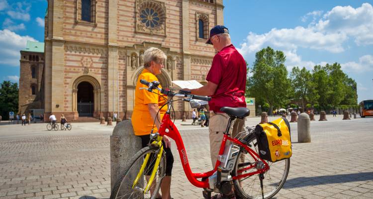 Ouder echtpaar bestudeert een kaart naast een tourfiets voor een imposante Romaanse kathedraal.