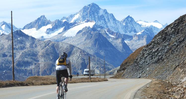 Un cycliste grimpe un col alpin sinueux vers des pics glaciaires imposants sous un ciel dégagé.