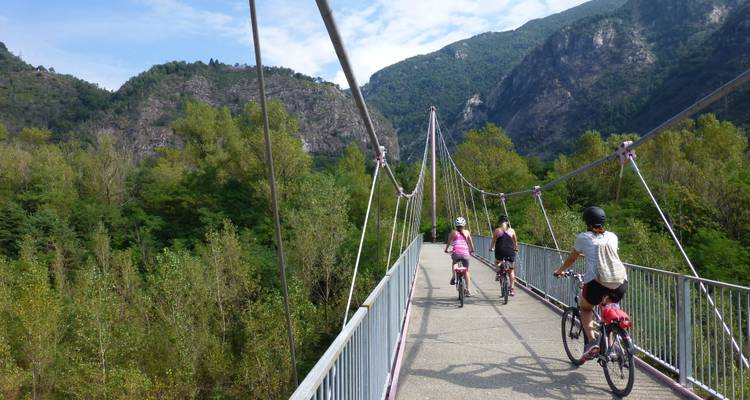 Cyclists crossing a suspension bridge with mountains in the background.