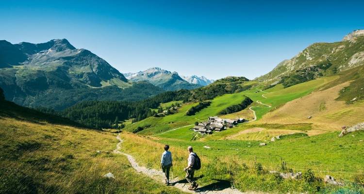 Deux randonneurs marchant sur un sentier de montagne avec des prairies vertes et des sommets en arrière-plan.