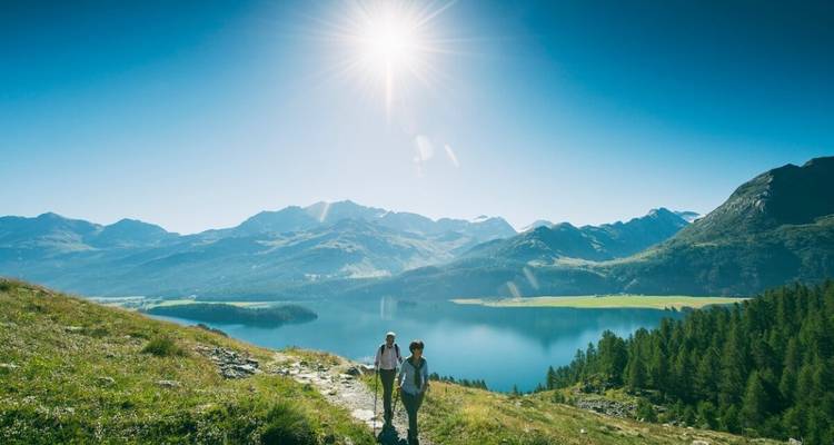Deux randonneurs sur un sentier ensoleillé surplombant un lac tranquille et un terrain montagneux.