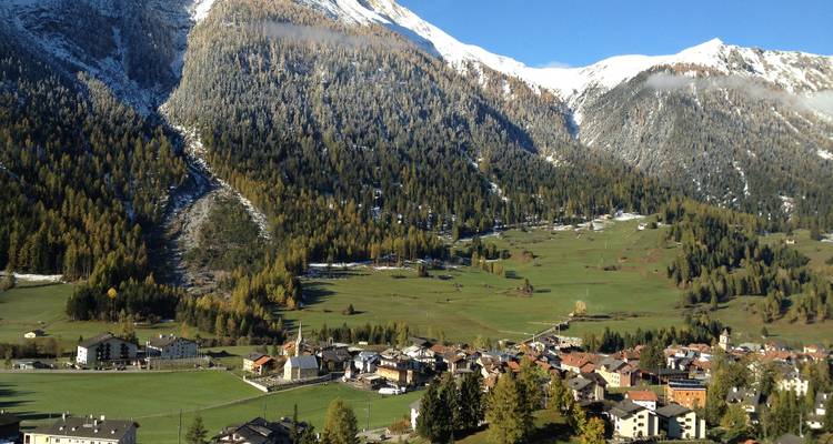 Alpine village nestled in a broad green valley beneath snow-dusted forests and peaks.
