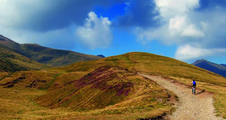 Ciclista en un sendero de tierra entre colinas onduladas bajo un cielo nublado.