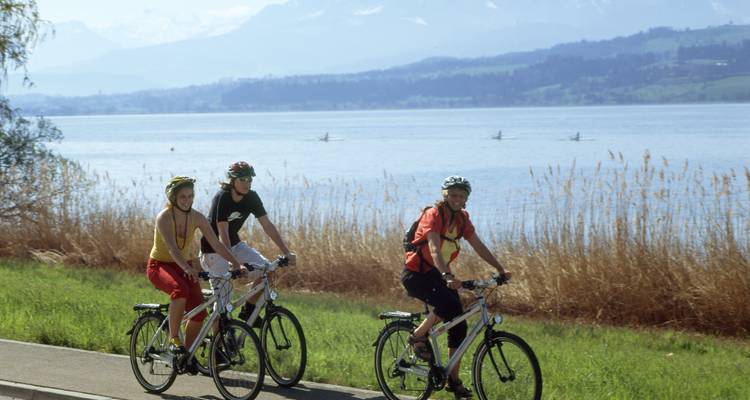 Ciclistas pedaleando junto a un lago con montañas de fondo.