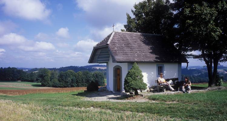 Small white roadside chapel in open farmland where two people sit on a bench enjoying the rural view.