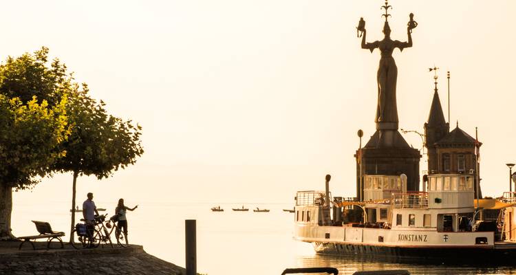 Eine Statue am Wasser mit einer Fähre im Hintergrund.