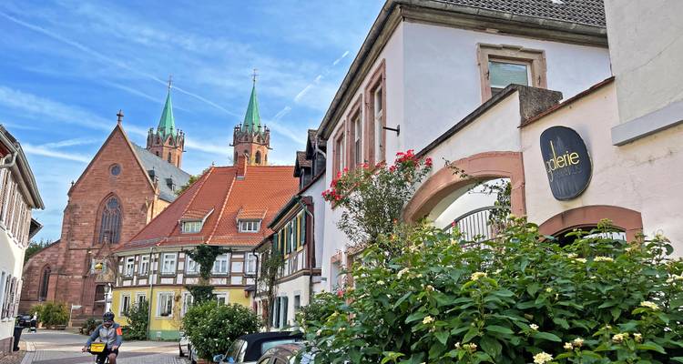 Quiet village street with colorful houses and twin church spires rising in the background on a bright day.