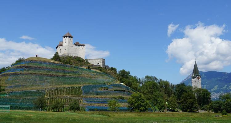 Château sur une colline verdoyante avec un clocher d'église au premier plan contre un ciel bleu.