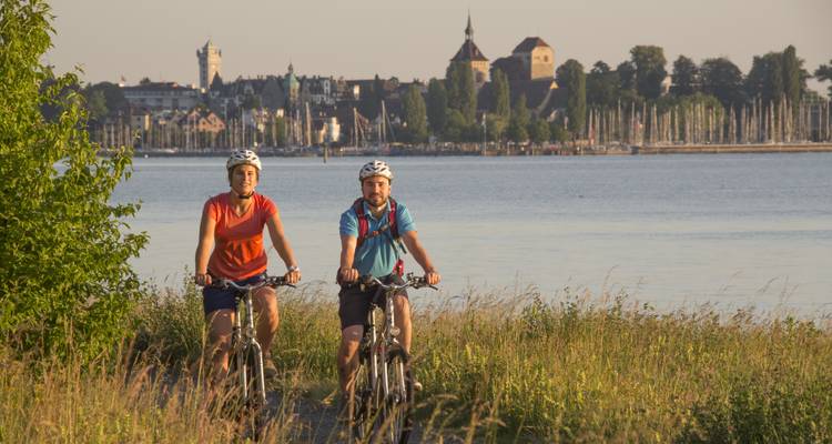 Deux cyclistes pédalant le long d'un sentier au bord d'un lac avec une vue sur une ville au loin.