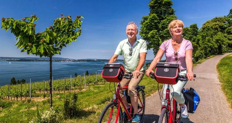 Ein Paar, das entlang eines malerischen Weges an einem See mit üppigem Grün radelt.