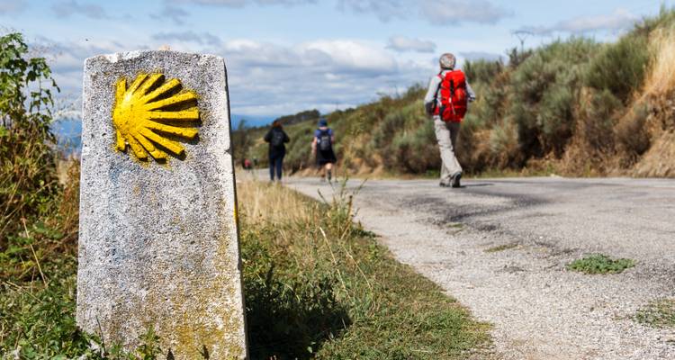 A stone marker with a yellow shell symbol and hikers in the distance.