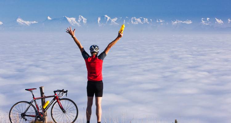 Radfahrer, der triumphierend die Arme in die Höhe streckt auf einer Bergstraße über den Wolken mit einer atemberaubenden Bergkette im Hintergrund.