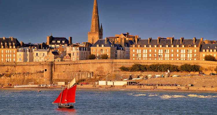 A sailboat with red sails in front of Saint-Malo city walls.