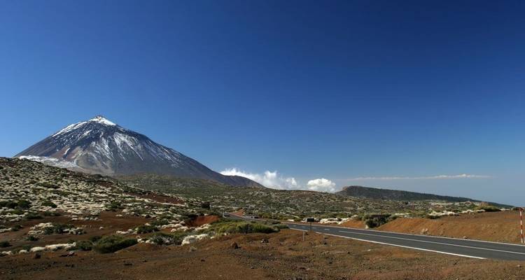 Route adjacente à un paysage volcanique brun-rouge avec un sommet enneigé au loin.