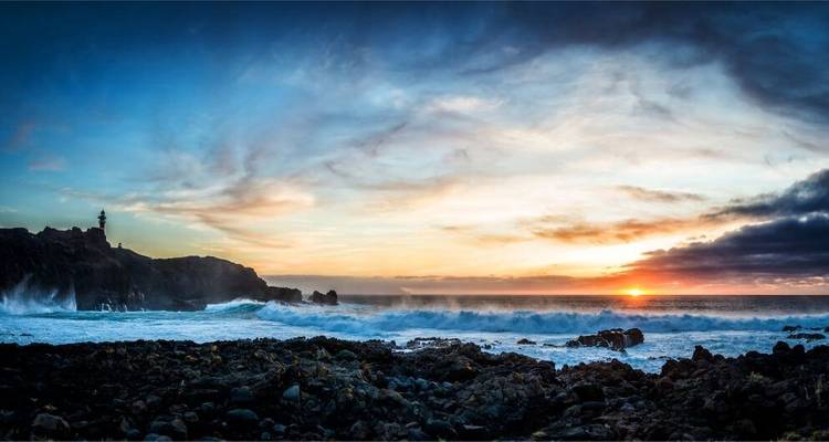 Sunset over rocky shoreline with waves crashing.