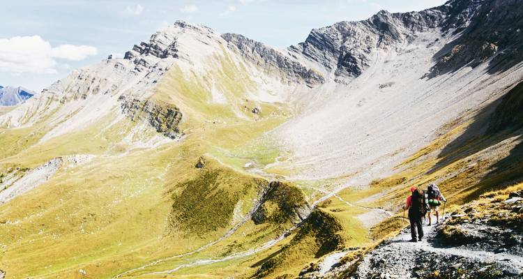 Zwei Wanderer wandeln entlang eines schmalen alpinen Pfades unter towering grauen Klippen und ausgedehnten U-förmigen Tälern.