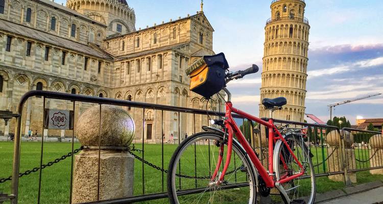 Een rode toerfiets staat bij een reling op Pisa's Piazza dei Miracoli met kathedraal en scheve toren in zonsondergangtinten.