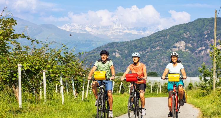 Trois cyclistes roulant sur un sentier pavé avec un lac bleu, une forêt et des montagnes alpines derrière eux.