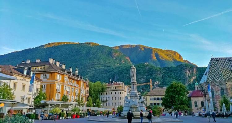 Una plaza animada en Bolzano con una estatua central, edificios amarillos y la luz dorada del atardecer resplandeciendo en las montañas circundantes.
