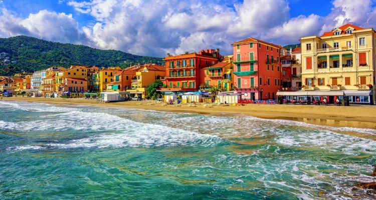 Lebendige ligurische Küstenstadt mit farbenfrohen Gebäuden entlang eines Sandstrandes und türkisfarbenen Wellen unter teilweise bewölktem Himmel.