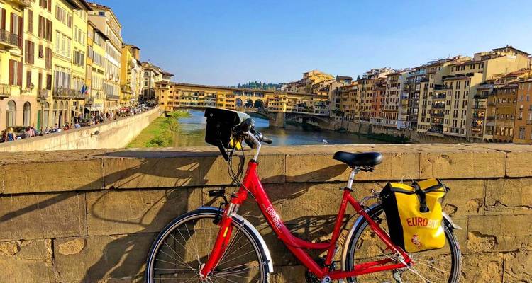 Een rode EuroBike leunt tegen een stenen brugmuur met uitzicht op de Ponte Vecchio van Florence en de rivier de Arno.