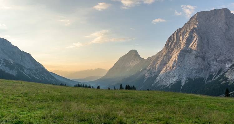 Weiches, goldenes Licht über einer ausgedehnten Alpwiese, die zu zerklüfteten Gipfeln und fernen, nebelverhangenen Tälern führt.