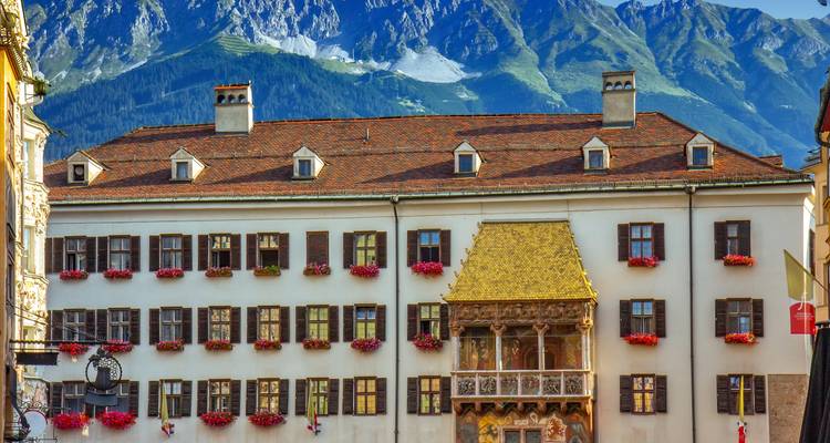 Historisches Gebäude mit goldenem Dach, geschmückt mit Geranien vor alpinen Bergen.