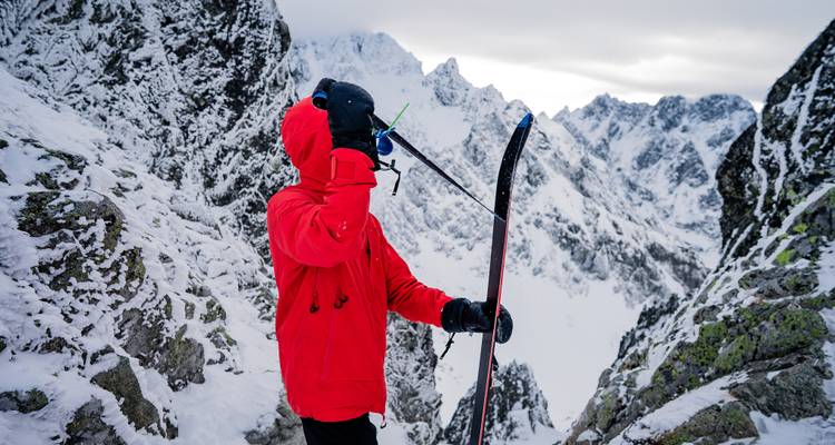 Skieur debout sur une crête rocheuse enneigée avec des montagnes en arrière-plan.