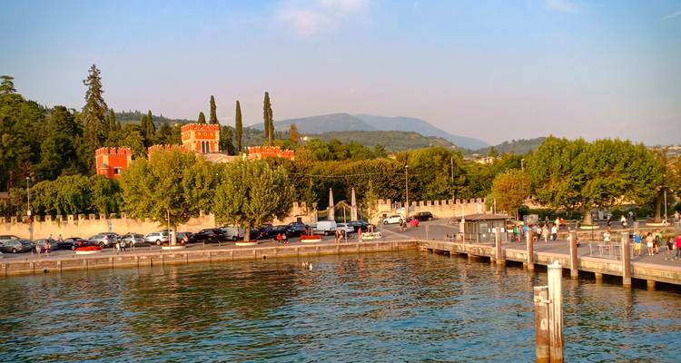 Vista desde el lago sobre un pequeño puerto, villa anaranjada tipo castillo y orilla arbolada bajo la cálida luz de la tarde.