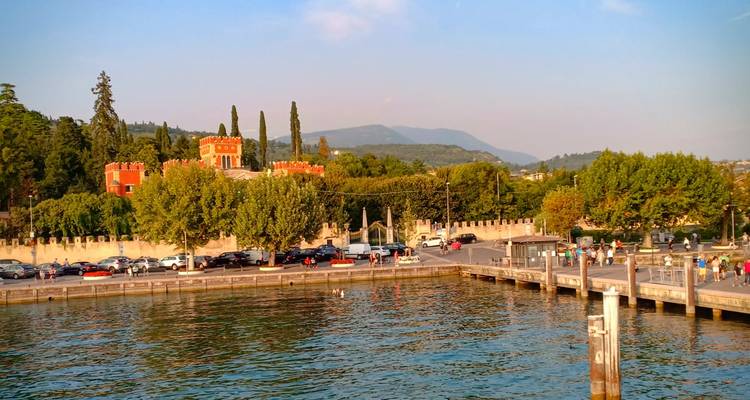 Uferpromenade mit burgähnlicher roter Villa, Pier und geparkten Autos am Ufer des Gardasees bei Sonnenuntergang