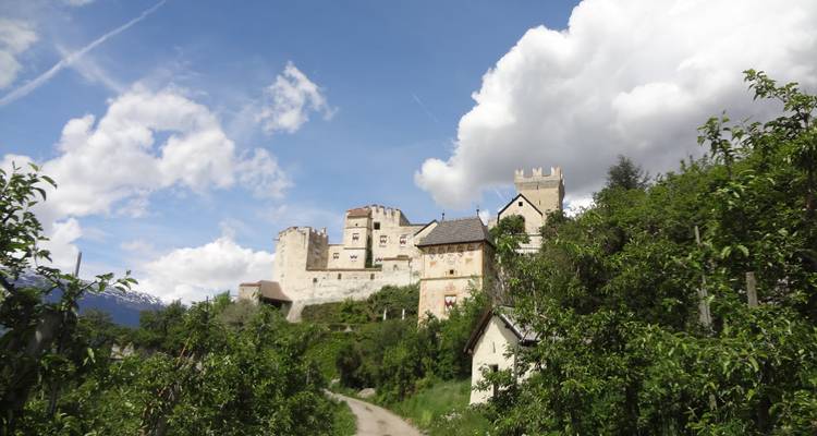 Castillo histórico en la cima de una colina rodeado de huertos y un sendero de tierra, con dramáticas nubes blancas en el cielo.