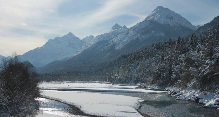 Montagnes enneigées et forêts le long d'une rivière avec un ciel nuageux.
