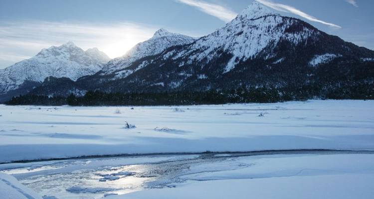 Paysage enneigé avec des montagnes et une rivière partiellement gelée sous un ciel ensoleillé.