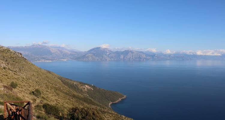 Weitläufige Küstenaussicht über ruhiges blaues Meer mit grünen Hügeln und fernen, dunstig verschleeierten Bergen.