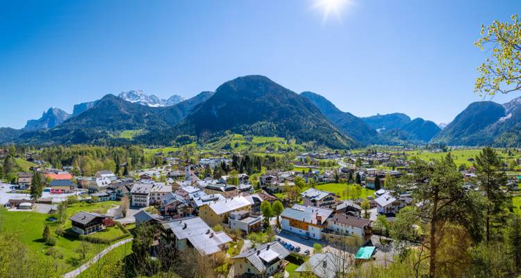 Panoramischer sonniger Blick über ein Alpendorf in einem grünen Tal, umgeben von dramatischen Bergen
