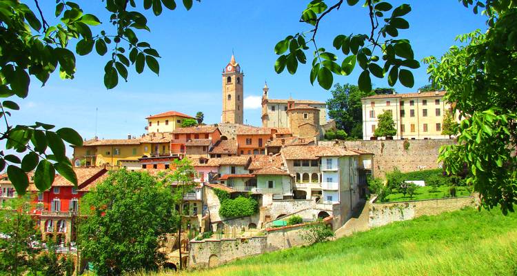 Village médiéval coloré avec une tour d'horloge proéminente vue à travers les branches d'arbres feuillues.