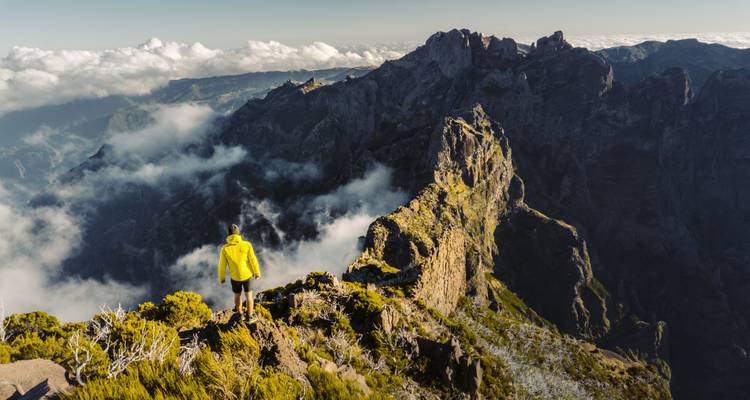 A person in a yellow jacket standing on a rocky mountain edge with clouds and peaks in the background.