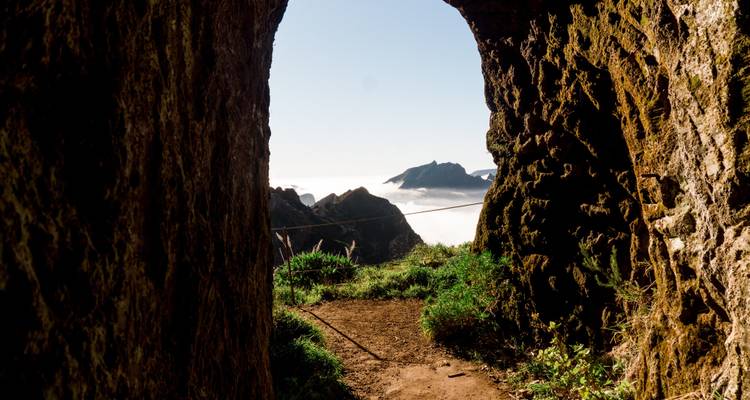 View through a rocky tunnel opening with mountains and clouds visible outside.