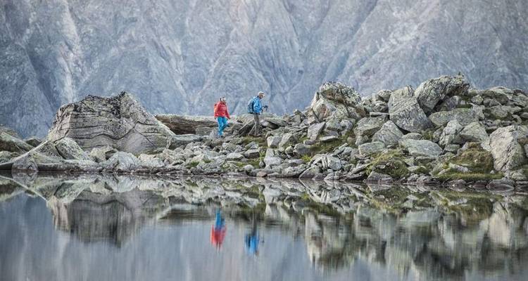 Deux personnes marchant sur un terrain rocheux au bord d'un lac calme et réfléchissant.