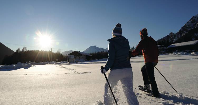 Dos personas caminando por un campo nevado con el sol poniéndose detrás de las montañas.