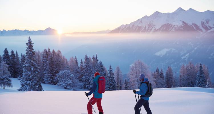 Dos personas caminando con raquetas de nieve al atardecer con una cordillera de fondo.