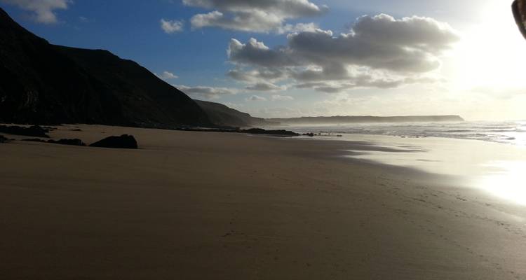 Plage de sable sombre avec des falaises et un ciel nuageux avec une faible luminosité solaire.