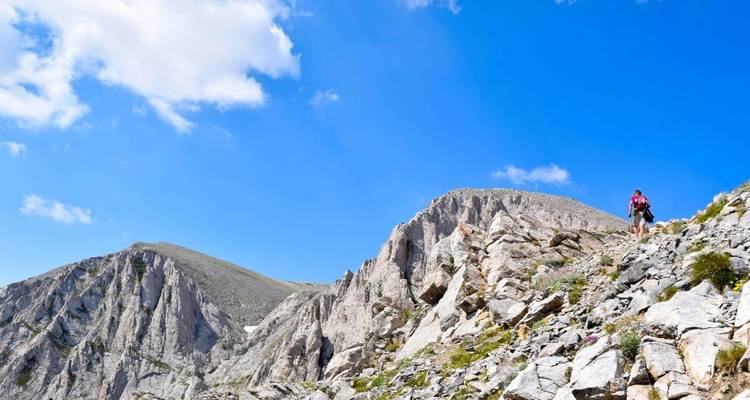 A hiker walking on a rocky mountain trail under a clear blue sky.