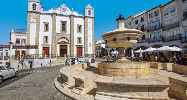 A lively square with a fountain and historical church, people are walking around.