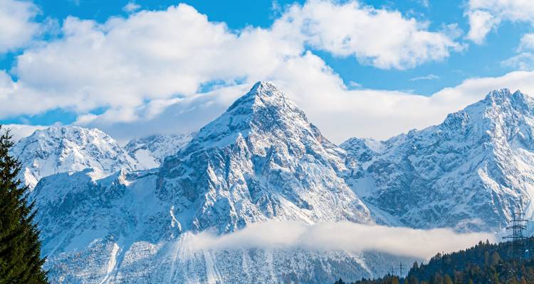 Sharp snow-capped Dolomite peak rising above clouds and evergreen forest in crystal clear light.