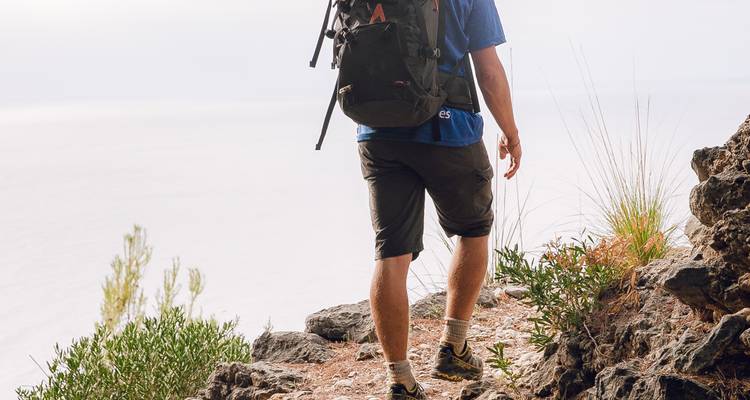 Gros plan des jambes et du sac à dos d'un randonneur sur un sentier rocheux.